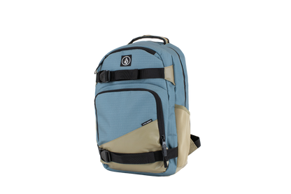The Volcom Men's Grindstone Skate Backpack is shown standing upright against a plain white background. It features a blue and beige color scheme with multiple zippered compartments, black ergonomic straps, a padded laptop sleeve, a top handle, and a circular Volcom logo patch on the front.