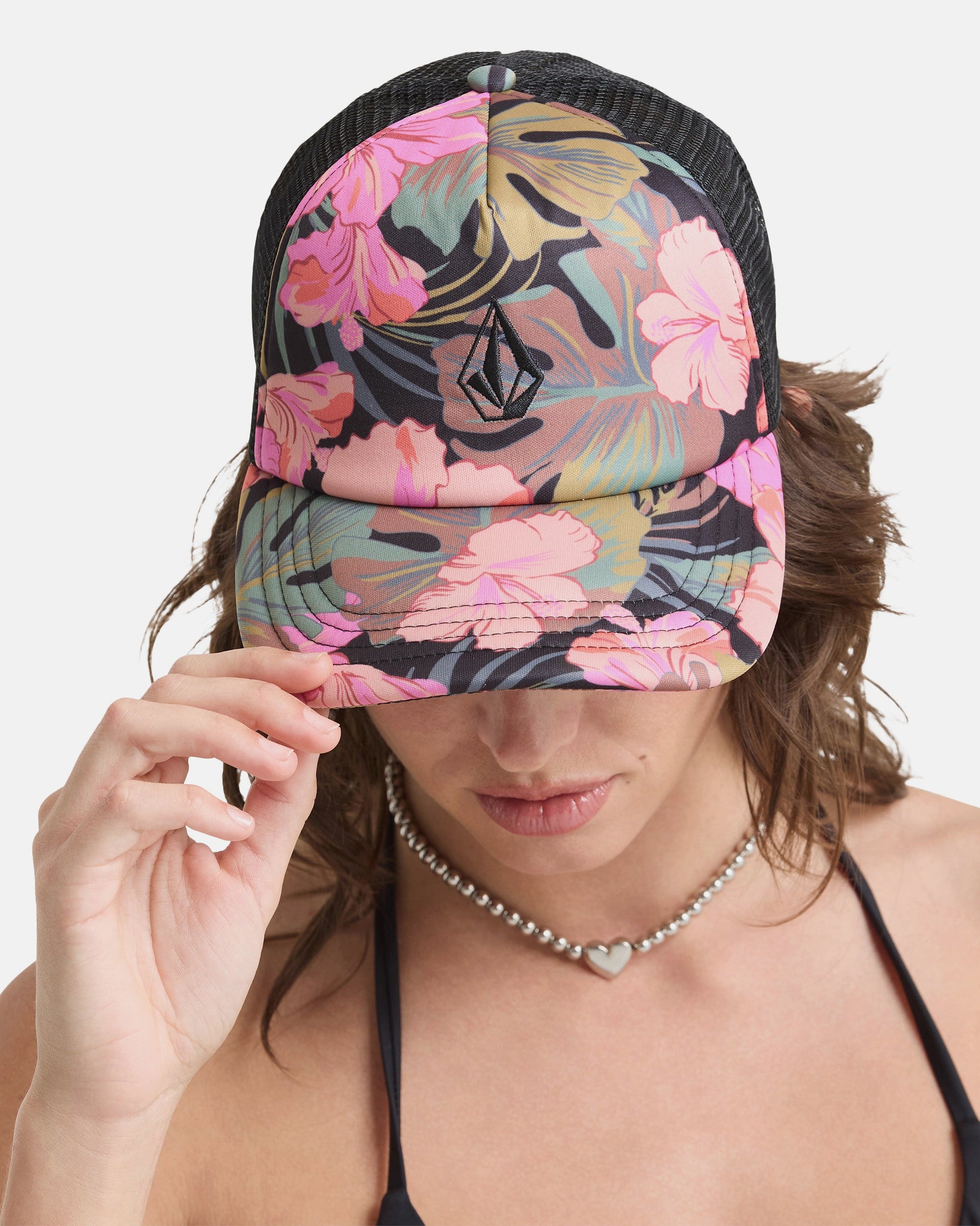 A person wearing the Volcom Women's Into Paradise Hat, featuring a black floral design with pink flowers, pulls the brim over their eyes. They pair it with a heart-shaped necklace and a black top.