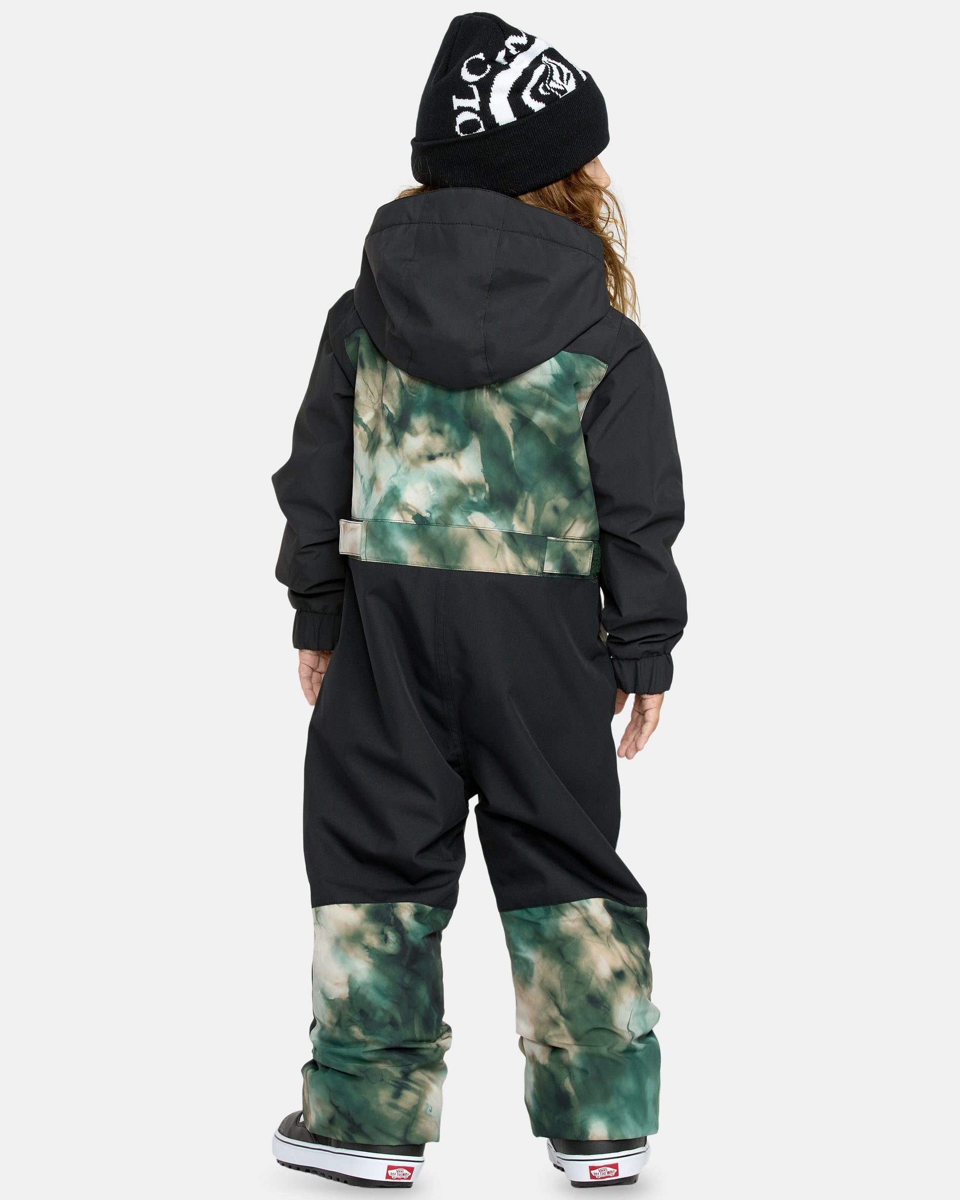 A child faces away, wearing the Kids' Little Flurry One Piece in black and green tie-dye, a black beanie with white lettering, and black-and-white sneakers, against a plain light background.