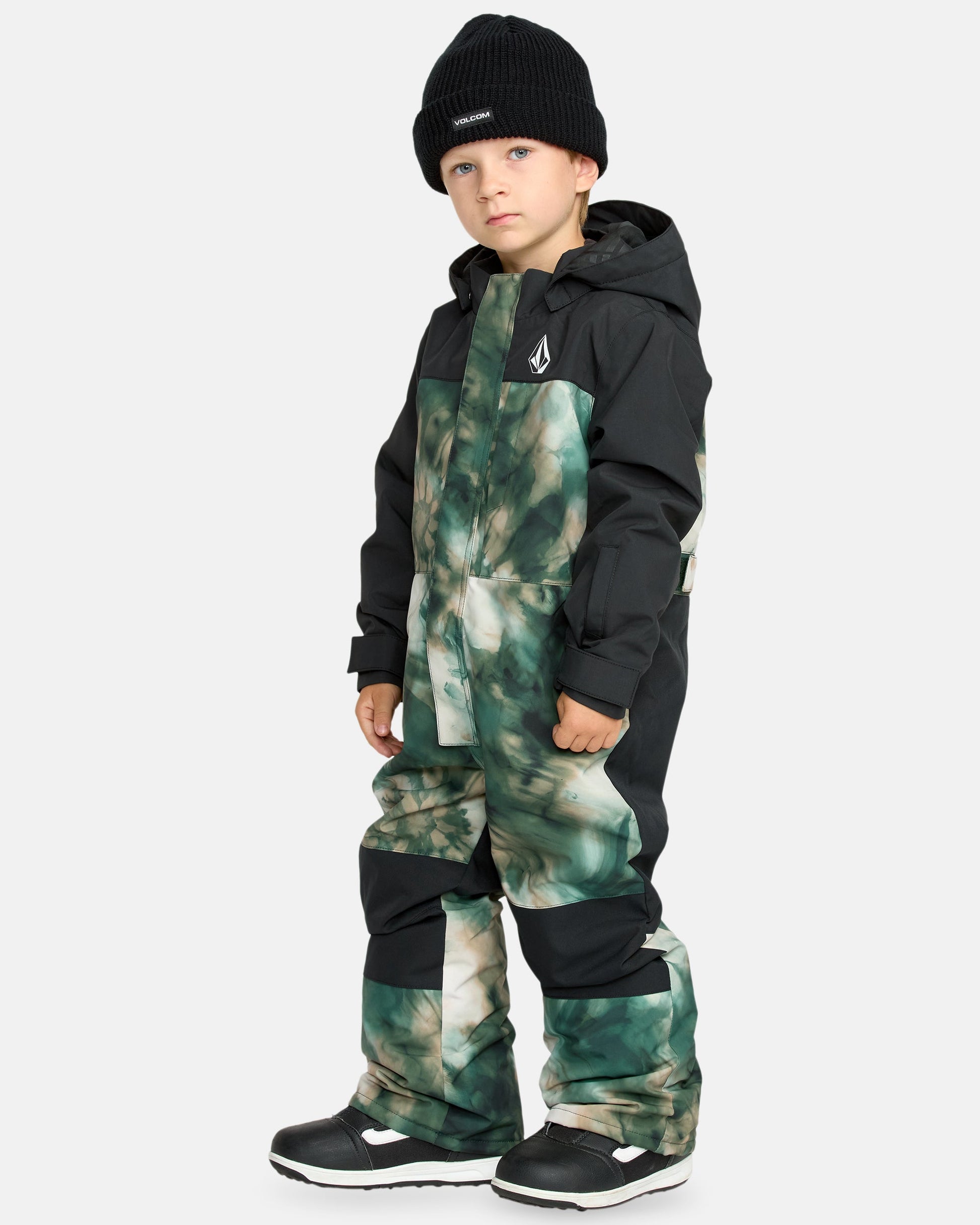 A young child stands facing forward in the Kids' Little Flurry One Piece—a hooded, green and black patterned snowsuit—paired with a black beanie and snow boots. The background is plain light gray.