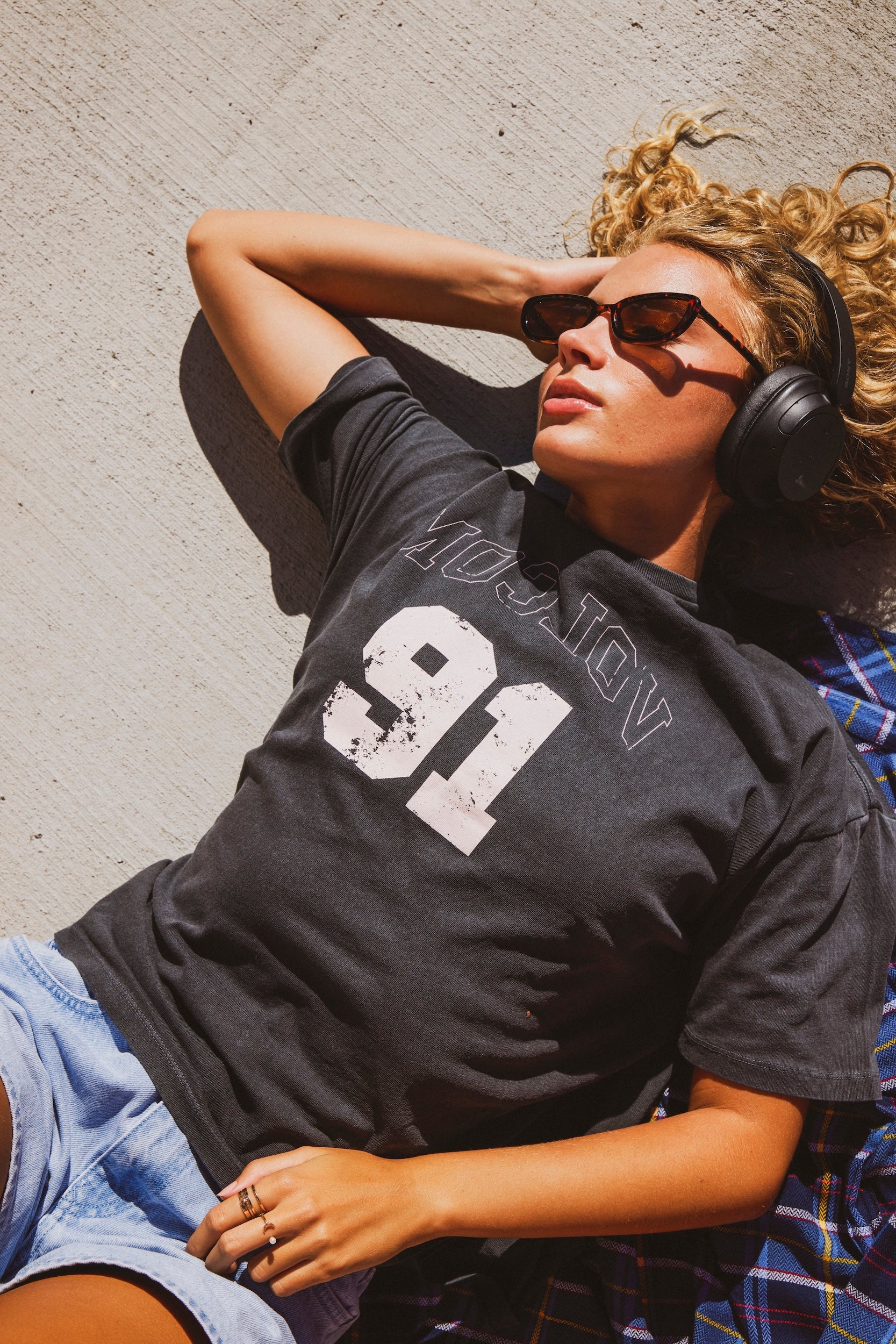 A woman wearing a black graphic t-shirt with a distressed white '91' and mirrored 'VOLCOM' text, lying on a concrete wall under bright sunlight. She is also wearing sunglasses, headphones, and denim shorts.