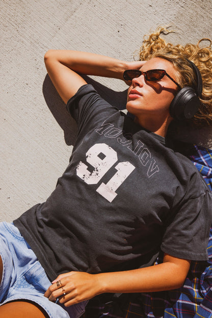 A woman wearing a black graphic t-shirt with a distressed white '91' and mirrored 'VOLCOM' text, lying on a concrete wall under bright sunlight. She is also wearing sunglasses, headphones, and denim shorts.